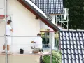 Dwóch malarzy na rusztowaniu odnawia elewację domu. Sprawdź, jaka jest malowanie elewacji cena.