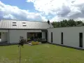 Nowoczesny dom z białymi ścianami i szarym stropodachem. Na tarasie stolik z krzesłami, obok żółta skrzynia z trawą.