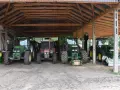 Ciężkie maszyny rolnicze, w tym zielone John Deere i czerwony Zetor, stoją pod drewnianą wiatą na działce rolnej, gotowe do pracy.