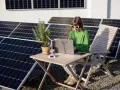 Dziewczyna w okularach pracuje na laptopie przy stole, otoczona panelami słonecznymi. Obok stoi model wiatraka, symbolizujący energię oze.
