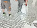 Pracownicy przygotowują wylanie posadzki z ogrzewaniem podłogowym. Układają białe rurki na siatce izolacyjnej.