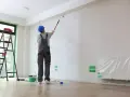 Malowanie ścian przez pracownika w niebieskim kasku. W tle drabina i wiadro z farbą. Określenie koszt malowania mieszkania.