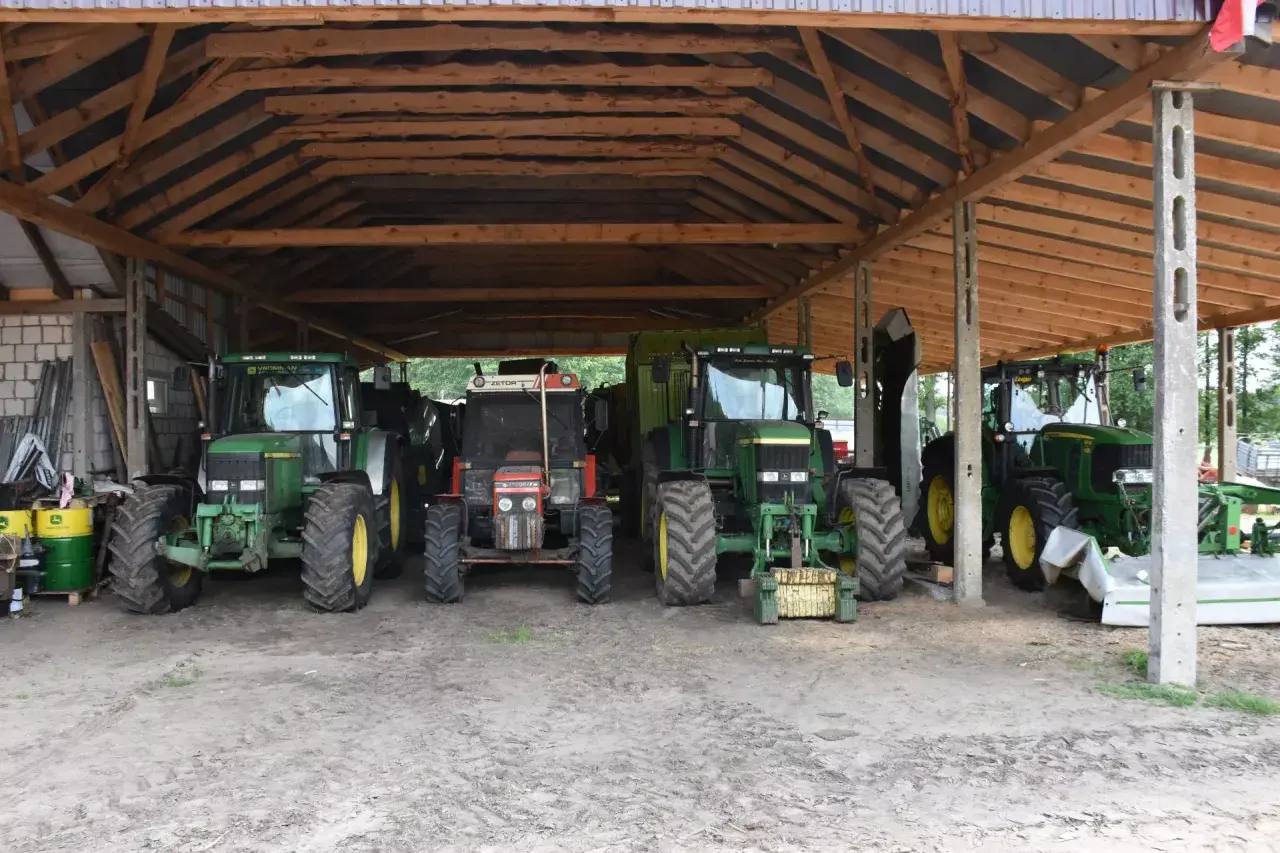 Ciężkie maszyny rolnicze, w tym zielone John Deere i czerwony Zetor, stoją pod drewnianą wiatą na działce rolnej, gotowe do pracy.