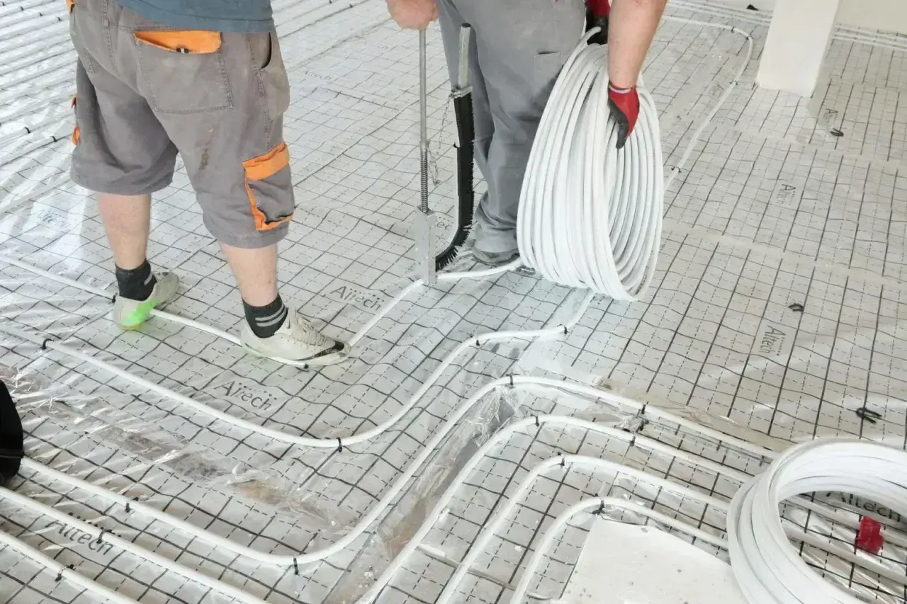 Pracownicy przygotowują wylanie posadzki z ogrzewaniem podłogowym. Układają białe rurki na siatce izolacyjnej.