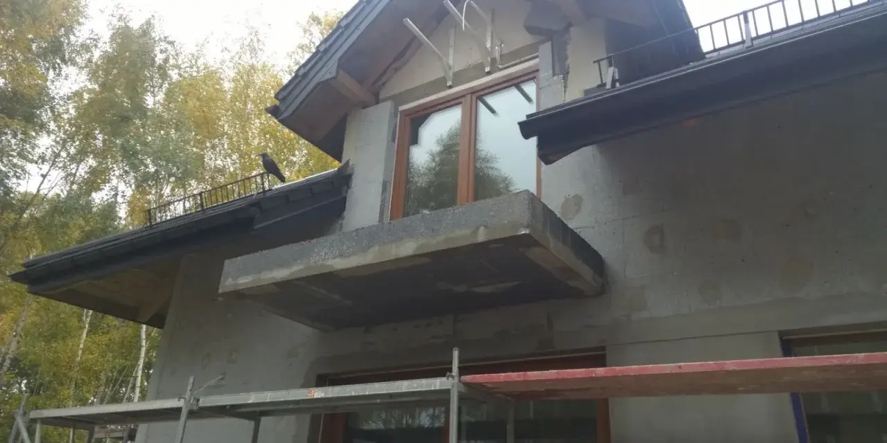Niedokończona elewacja domu z balkonem i oknem. Na dachu siedzi ptak.