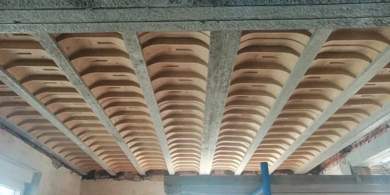 Widok od dołu na strop w domu parterowym, z widocznymi belkami stropowymi i pustakami ceramicznymi.