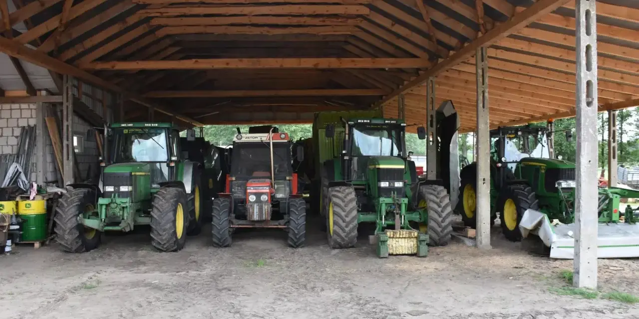 Ciężkie maszyny rolnicze, w tym zielone John Deere i czerwony Zetor, stoją pod drewnianą wiatą na działce rolnej, gotowe do pracy.