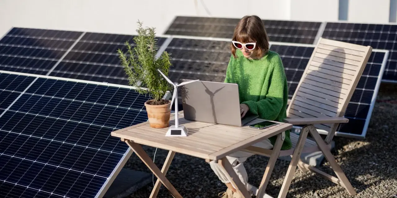 Dziewczyna w okularach pracuje na laptopie przy stole, otoczona panelami słonecznymi. Obok stoi model wiatraka, symbolizujący energię oze.