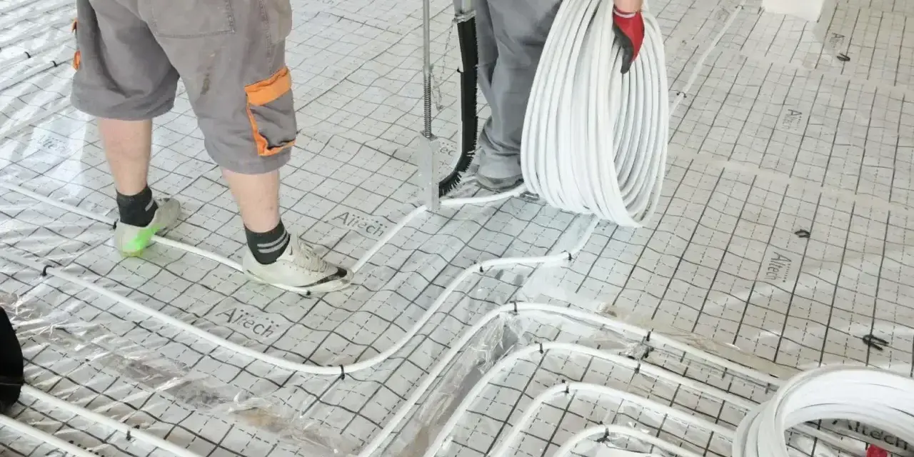 Pracownicy przygotowują wylanie posadzki z ogrzewaniem podłogowym. Układają białe rurki na siatce izolacyjnej.