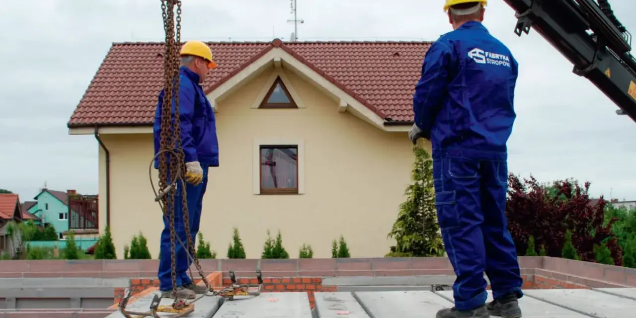Dwóch budowlańców w żółtych kaskach i niebieskich kombinezonach montuje prefabrykowany strop. Jeden z nich stoi na płycie, drugi czeka na dalsze instrukcje.