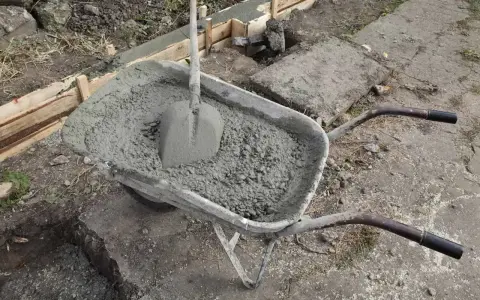 Taczka z betonem i łopatą czeka na użycie przy budowie fundamentów. W tle widać szalunki i przygotowane zbrojenie.