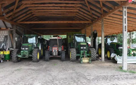 Ciężkie maszyny rolnicze, w tym zielone John Deere i czerwony Zetor, stoją pod drewnianą wiatą na działce rolnej, gotowe do pracy.