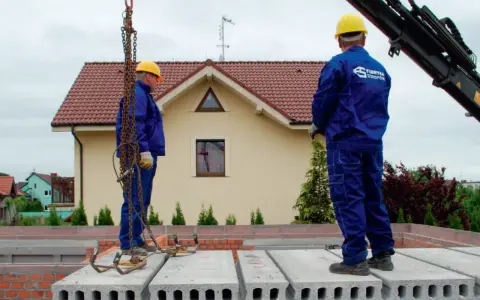 Dwóch budowlańców w żółtych kaskach i niebieskich kombinezonach montuje prefabrykowany strop. Jeden z nich stoi na płycie, drugi czeka na dalsze instrukcje.