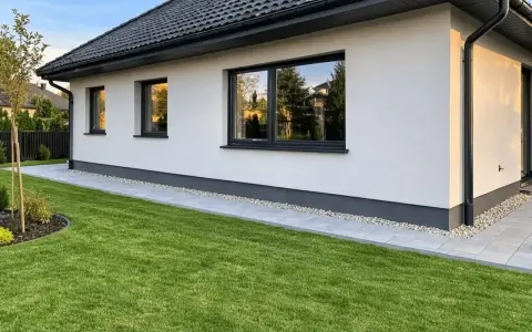 Nowy dom z białym tynkiem i czarnym dachem. Wokół domu ścieżka z płyt i zielona trawa. To najtańsza opaska wokół domu, która dodaje uroku.