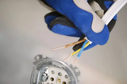 Rękawica pokazuje, jak podłączyć gniazdko elektryczne. Widoczne przewody: brązowy, niebieski i żółto-zielony.