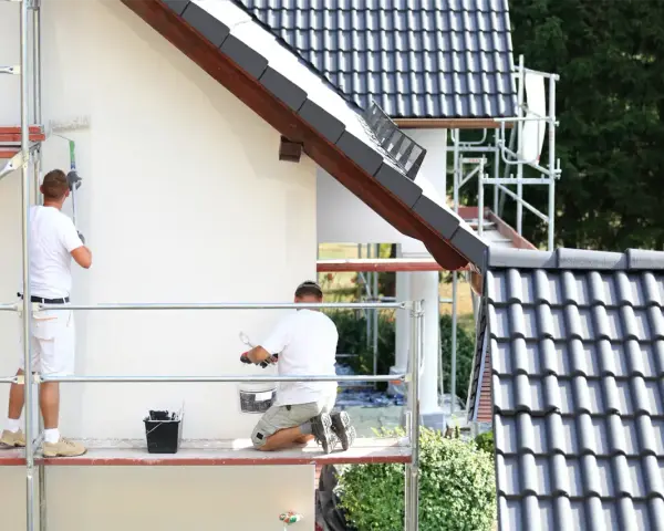 Dwóch malarzy na rusztowaniu odnawia elewację domu. Sprawdź, jaka jest malowanie elewacji cena.