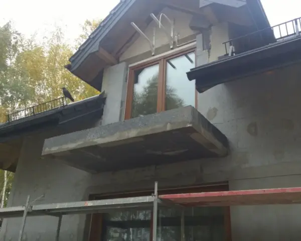 Niedokończona elewacja domu z balkonem i oknem. Na dachu siedzi ptak.