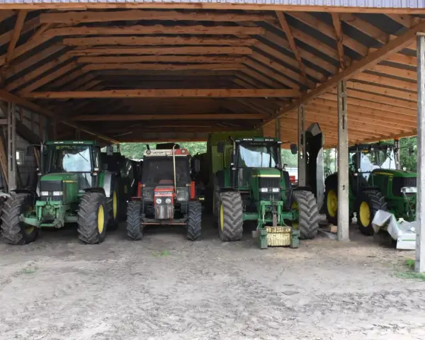 Ciężkie maszyny rolnicze, w tym zielone John Deere i czerwony Zetor, stoją pod drewnianą wiatą na działce rolnej, gotowe do pracy.