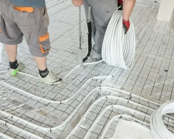 Pracownicy przygotowują wylanie posadzki z ogrzewaniem podłogowym. Układają białe rurki na siatce izolacyjnej.