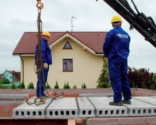 Dwóch budowlańców w żółtych kaskach i niebieskich kombinezonach montuje prefabrykowany strop. Jeden z nich stoi na płycie, drugi czeka na dalsze instrukcje.