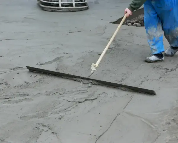 Pracownik w niebieskich spodniach wykonuje protokół wygrzewania posadzki, wygładzając świeży beton długą łopatą.
