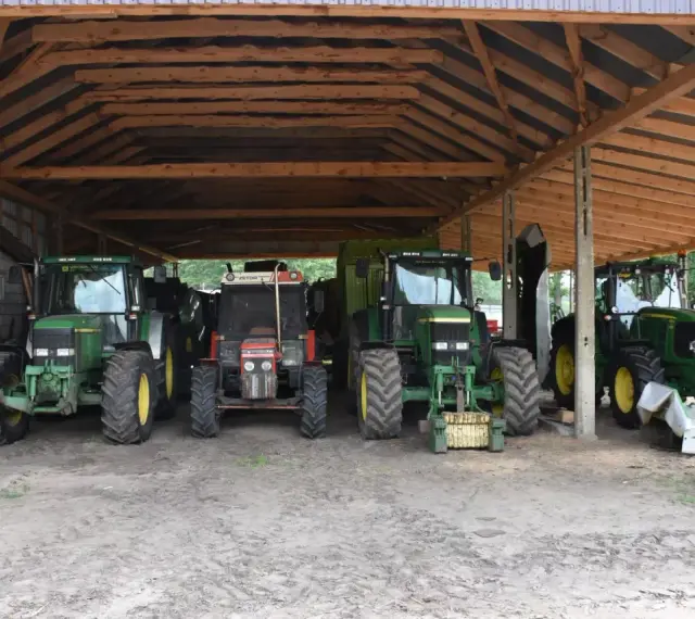 Ciężkie maszyny rolnicze, w tym zielone John Deere i czerwony Zetor, stoją pod drewnianą wiatą na działce rolnej, gotowe do pracy.