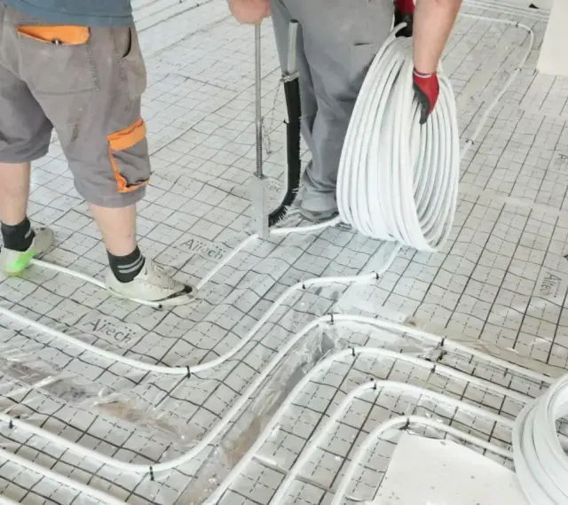 Pracownicy przygotowują wylanie posadzki z ogrzewaniem podłogowym. Układają białe rurki na siatce izolacyjnej.