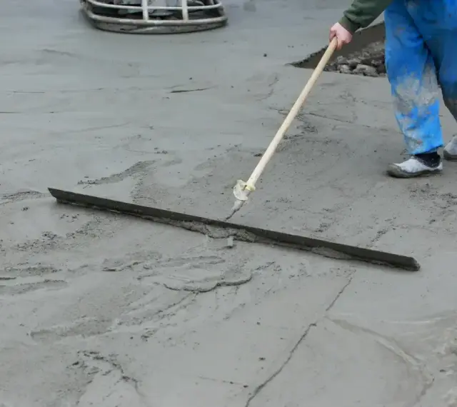 Pracownik w niebieskich spodniach wykonuje protokół wygrzewania posadzki, wygładzając świeży beton długą łopatą.
