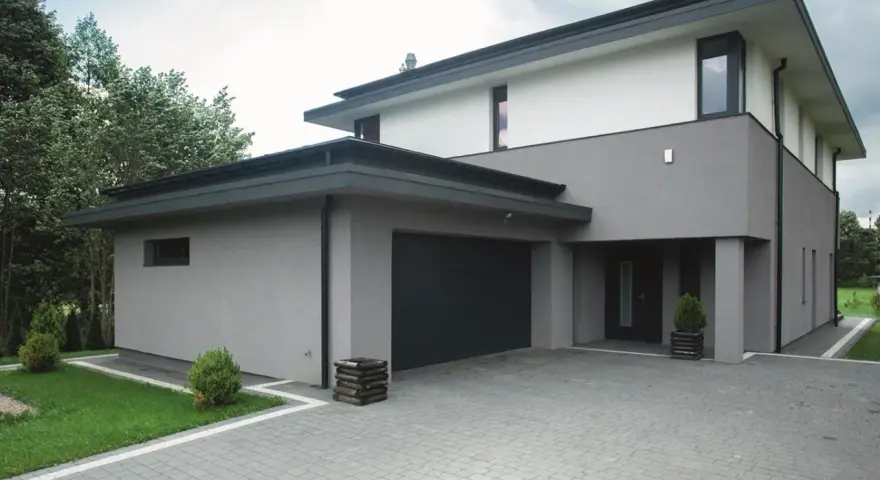 Nowoczesny dom z szarymi i białymi tynkami, z ciemnym dachem i bramą garażową. Eleganckie kolory tynków na domach tworzą minimalistyczną całość.