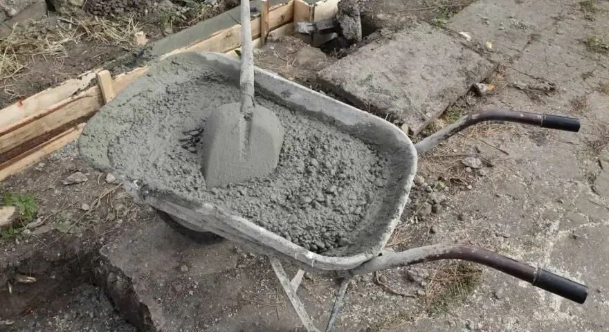 Taczka z betonem i łopatą czeka na użycie przy budowie fundamentów. W tle widać szalunki i przygotowane zbrojenie.