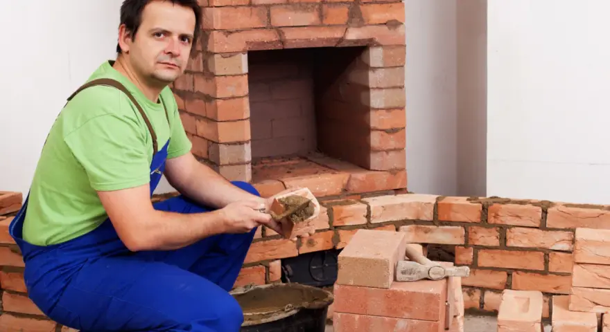 Murarz układa cegły szamotowe, budując kominek. W wiadrze zaprawa, obok stosy cegieł.