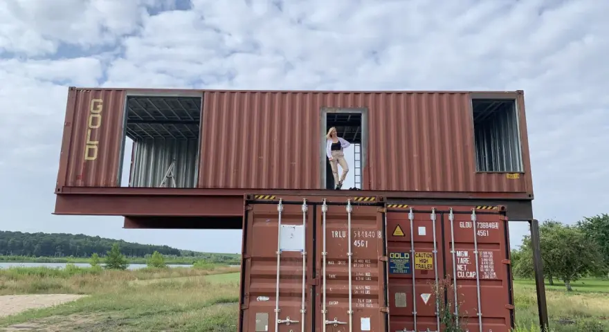 Kobieta stoi w drzwiach domu z kontenera, który jest częścią dwupoziomowej konstrukcji.