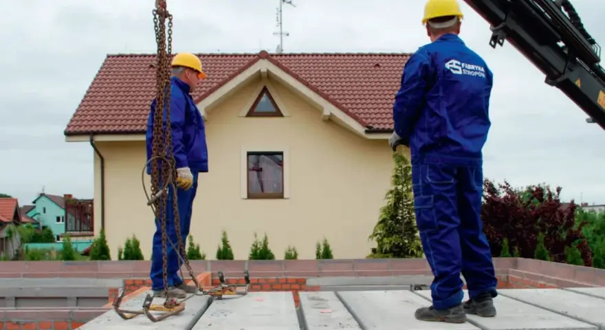 Dwóch budowlańców w żółtych kaskach i niebieskich kombinezonach montuje prefabrykowany strop. Jeden z nich stoi na płycie, drugi czeka na dalsze instrukcje.