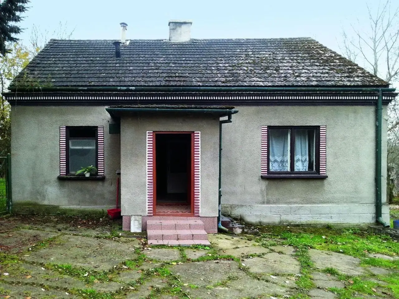 Rozpoczęty remont starego domu na wsi. Szary tynk, dach pokryty mchem, nowe okna i drzwi czekają na odnowienie.
