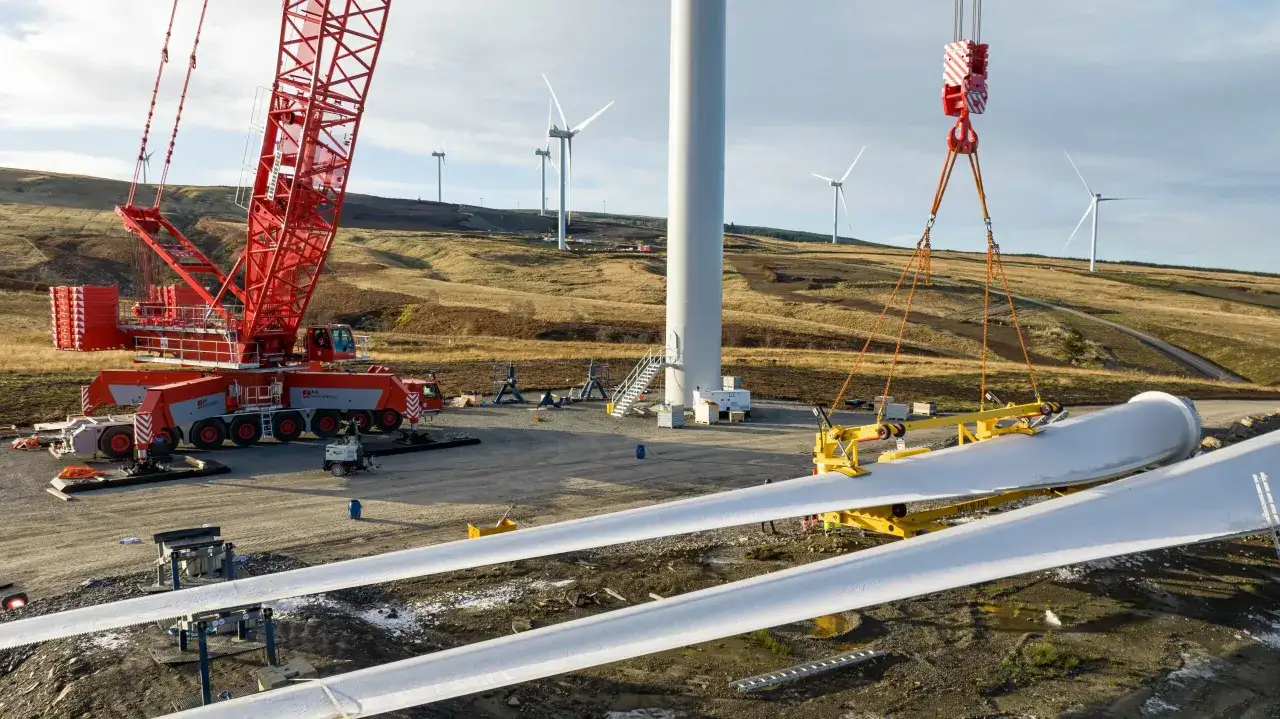 Gigantyczny dźwig montuje łopatę elektrowni wiatrowej na tle innych turbin na wzgórzu.
