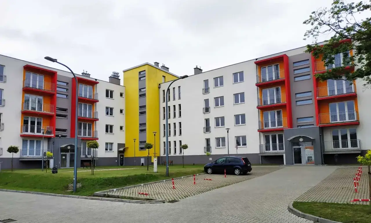 Nowoczesne bloki mieszkalne z kolorowymi balkonami, przypominające cegłę żerańską, otoczone zielenią i parkingiem.
