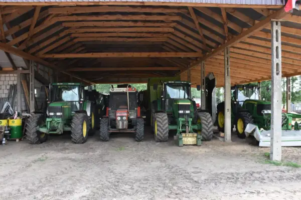 Ciężkie maszyny rolnicze, w tym zielone John Deere i czerwony Zetor, stoją pod drewnianą wiatą na działce rolnej, gotowe do pracy.