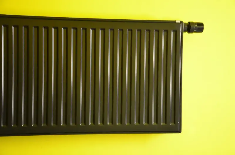 Nowoczesne malowanie grzejnika aluminiowego na ciemny kolor. Grzejnik z termostatem na żółtej ścianie.